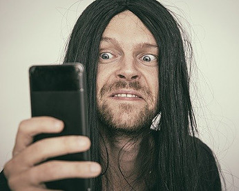 Shocked looking man with long black hair staring worriedly at his phone. 