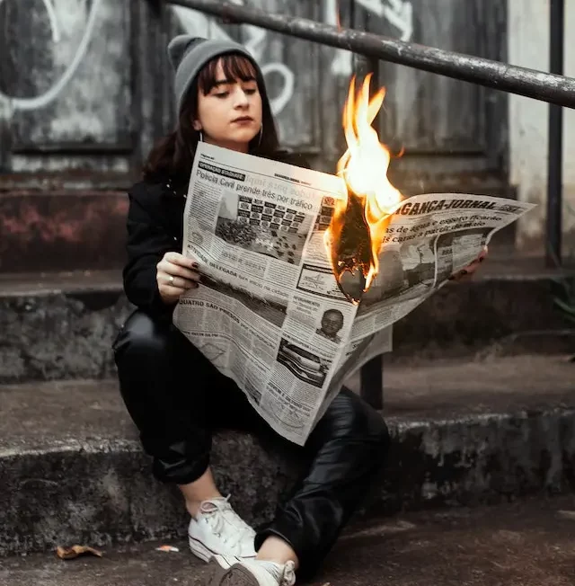 Woman sitting on the street reading a newspaper which is on fire.