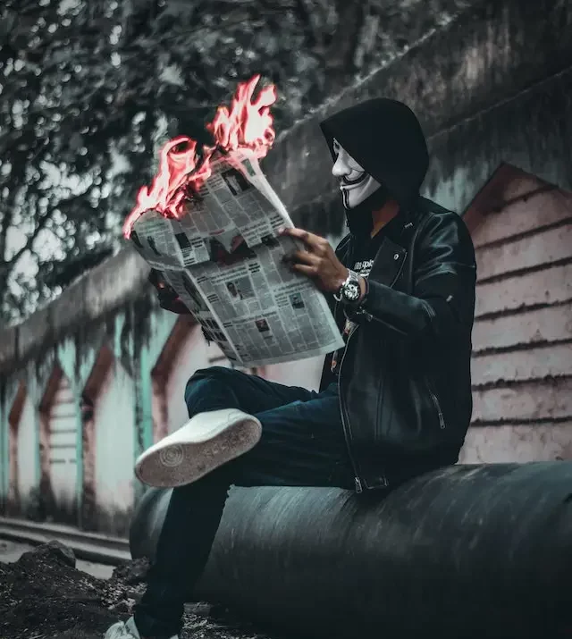 Masked man reading a newspaper which is on fire. 