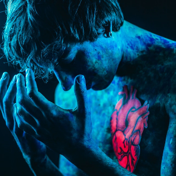 A man bathed in blue light with head in hands, a red heart on his chest. 