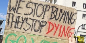 A sign held high at a protest reading "We stop buying, they stop dying - go vegan".