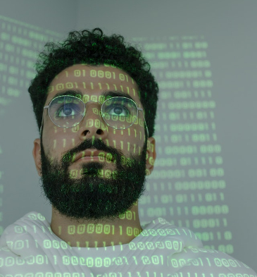 Neatly bearded man looking at a screen with a green projection of zeros and ones onto him.