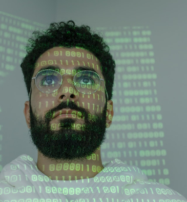 Neatly bearded man looking at a screen with a green projection of zeros and ones onto him. 