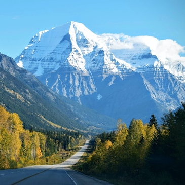 A road leading to the base of a tall mountain with snowy peaks and clouds drifting across the top. 