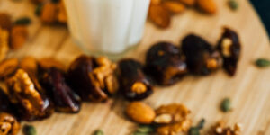 A glass of almond milk on a wooden chopping board scattered with almonds, other nuts and seeds with some dates on there too.