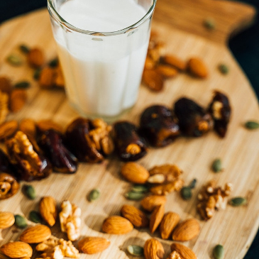 A glass of almond milk on a wooden chopping board scattered with almonds, other nuts and seeds with some dates on there too. 