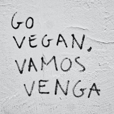 Grafitti on a white wall with flaking paint , saying "Go Vegan, Vamos Venga". 